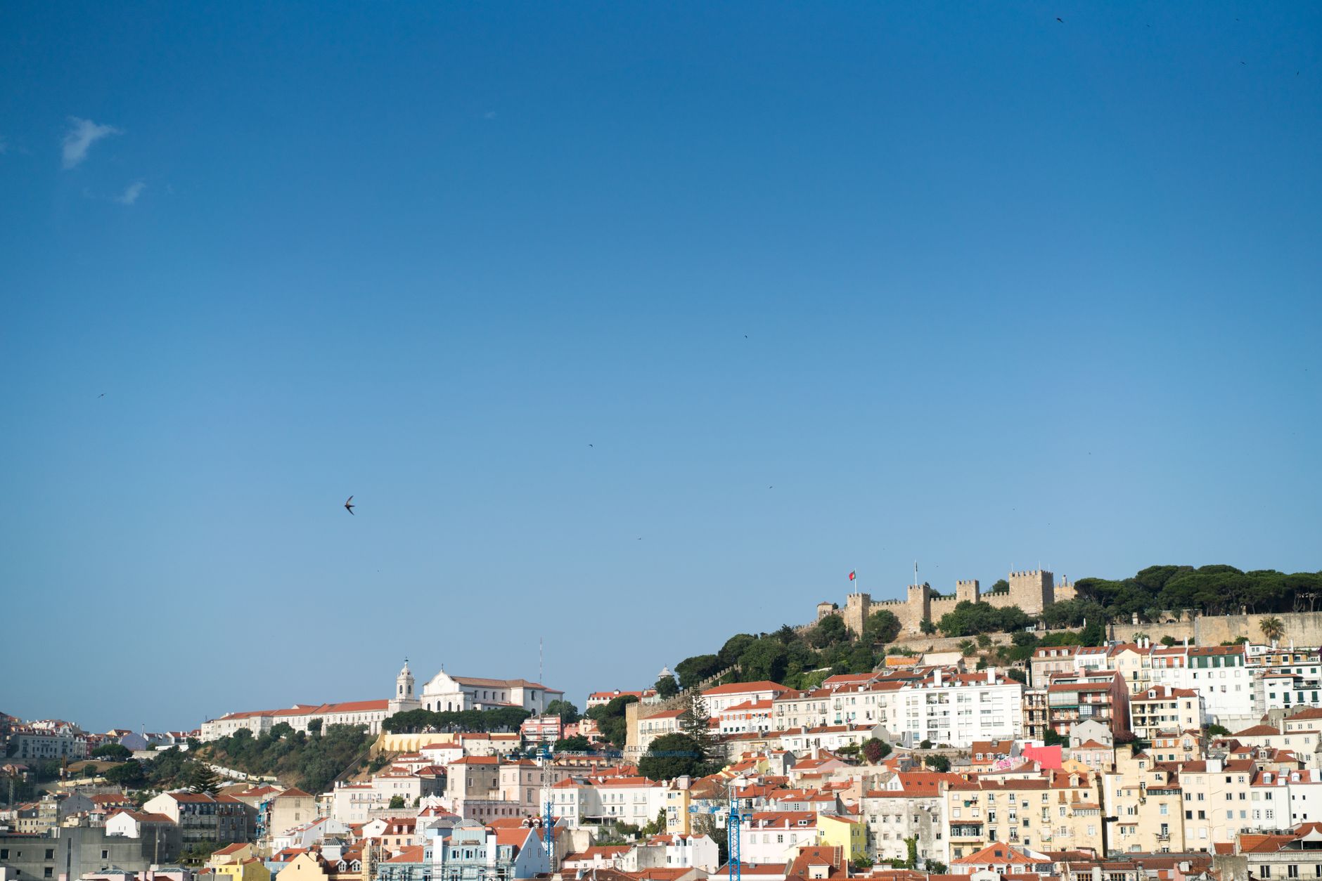 Panoramic view of Lisbon's historic center with terracotta rooftops, a winding river, and a mix of ancient and modern buildings under a clear sky.