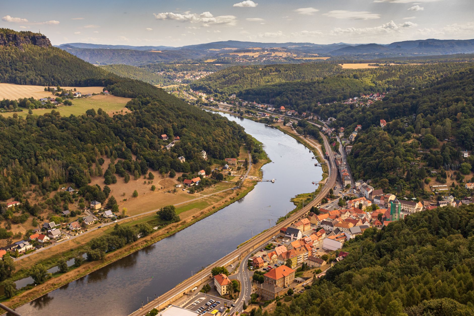 A sweeping aerial view of a picturesque German village nestled in rolling hills, with traditional houses and a church steeple, under a clear sky.