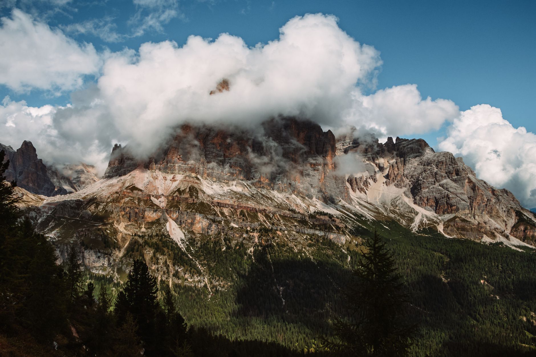 Sweeping view of the Dolomites in summer, showing vast green meadows, jagged grey peaks under a blue sky, with a hiking trail winding through the foreground.