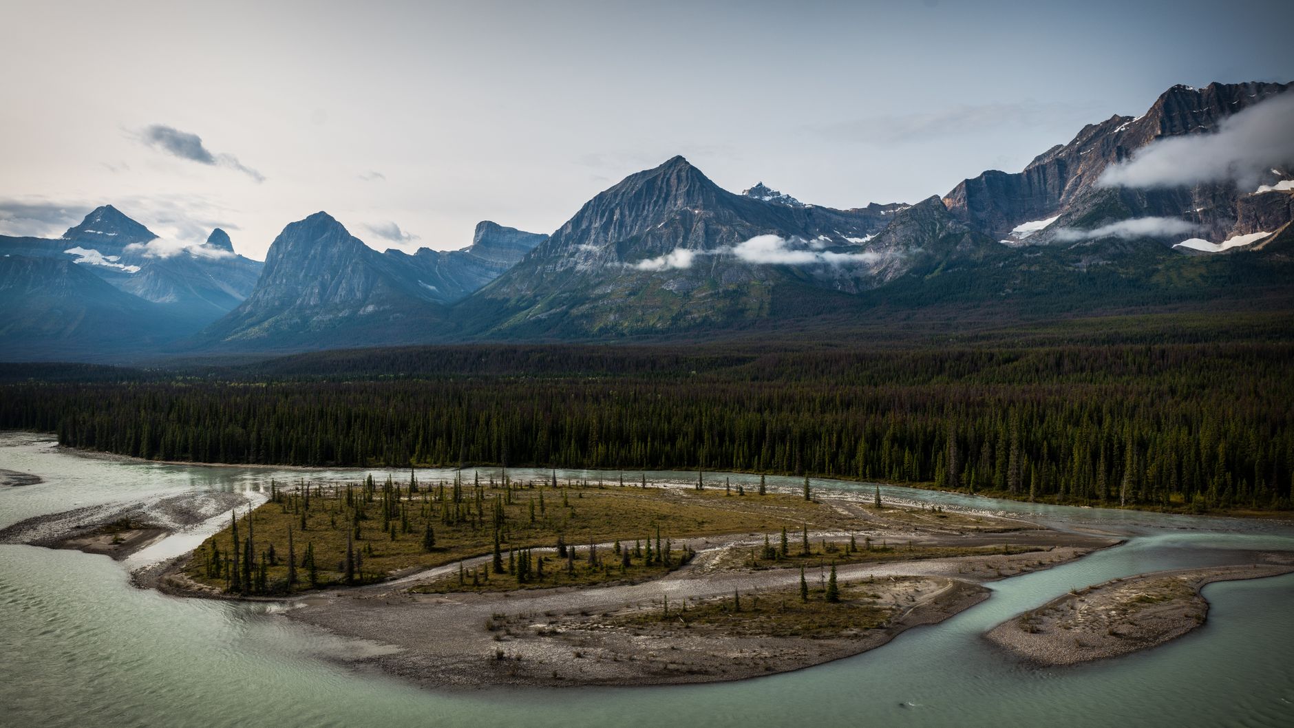Sweeping view of a vast Canadian wilderness, featuring towering mountains, dense forests, and a pristine lake under a clear sky.
