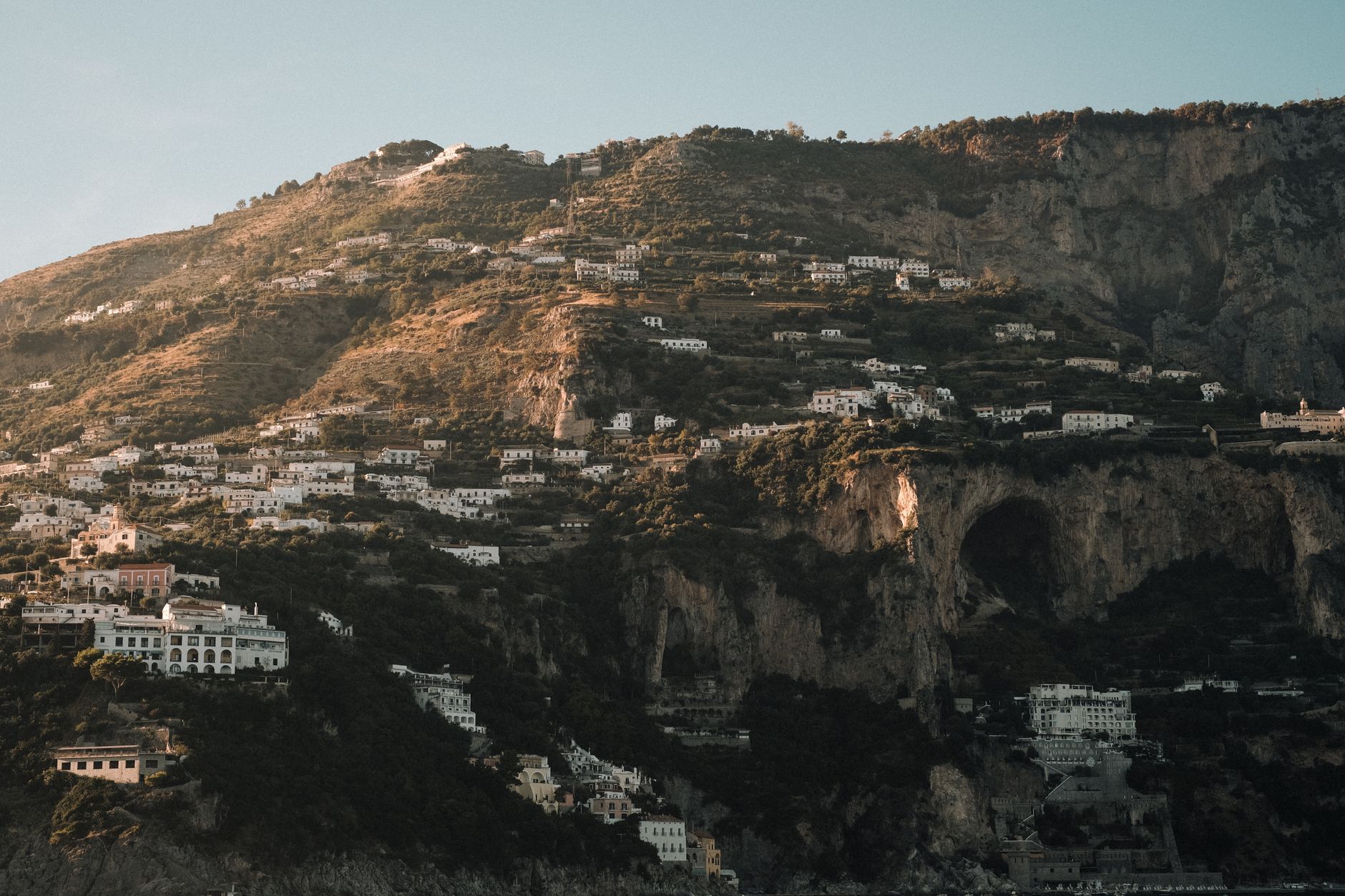A panoramic view of the rugged Amalfi Coast coastline, showing dramatic cliffs, azure waters, and a cluster of colorful, lesser-known villages clinging to the hillsides under a clear sky.
