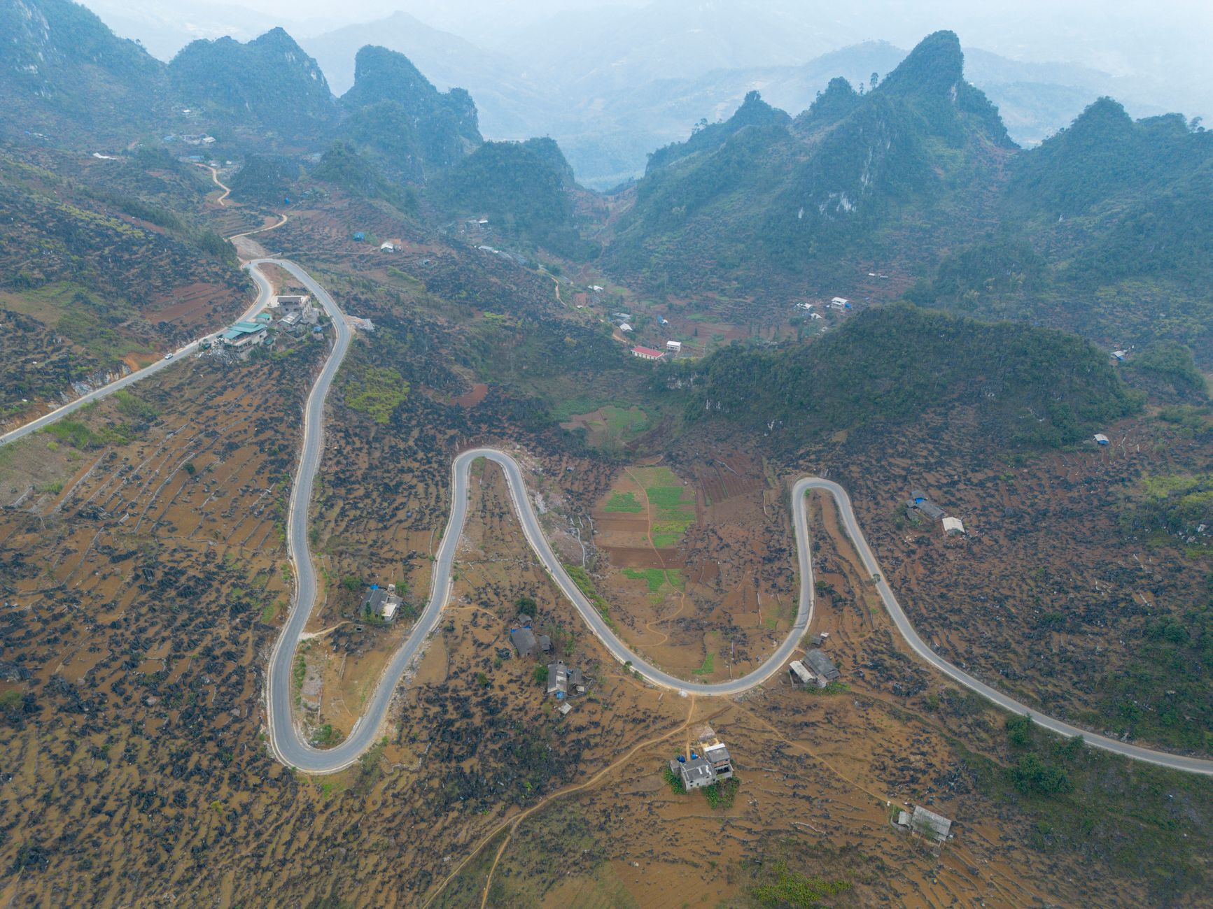 Sweeping aerial view of lush green limestone karsts shrouded in mist, with a winding road snaking through the valleys, characteristic of northern Vietnam's remote landscapes.