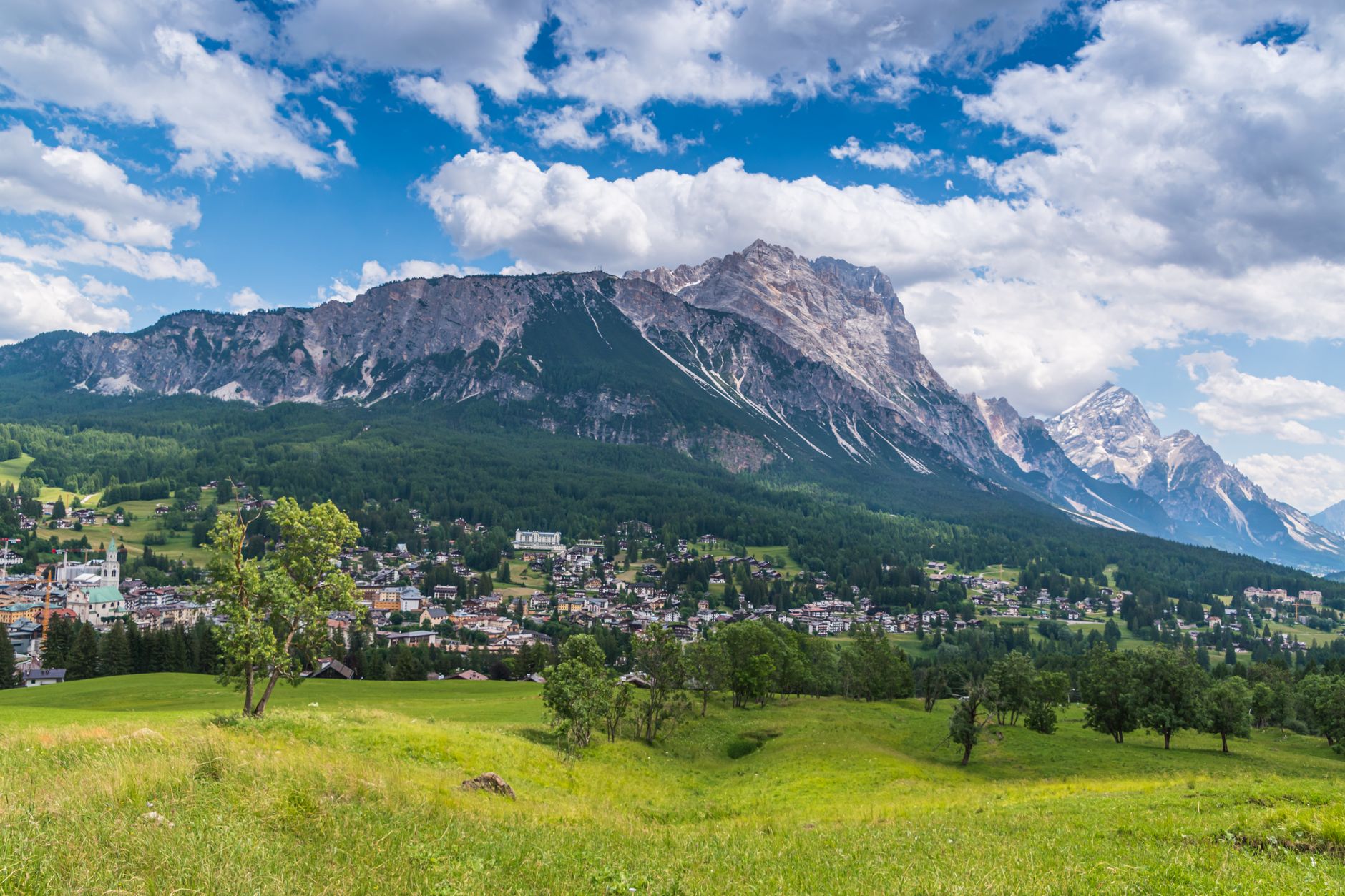 Sweeping panoramic view of a charming alpine village nestled in a green valley, surrounded by towering, jagged Dolomite peaks under a clear sky.