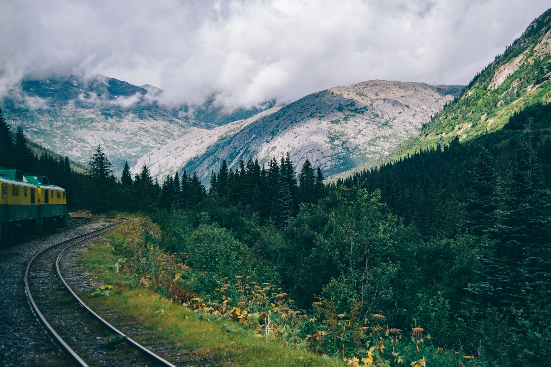 A sweeping landscape showing both an old train track curving through a lush valley and a person cycling on a path alongside a river, symbolizing diverse travel.