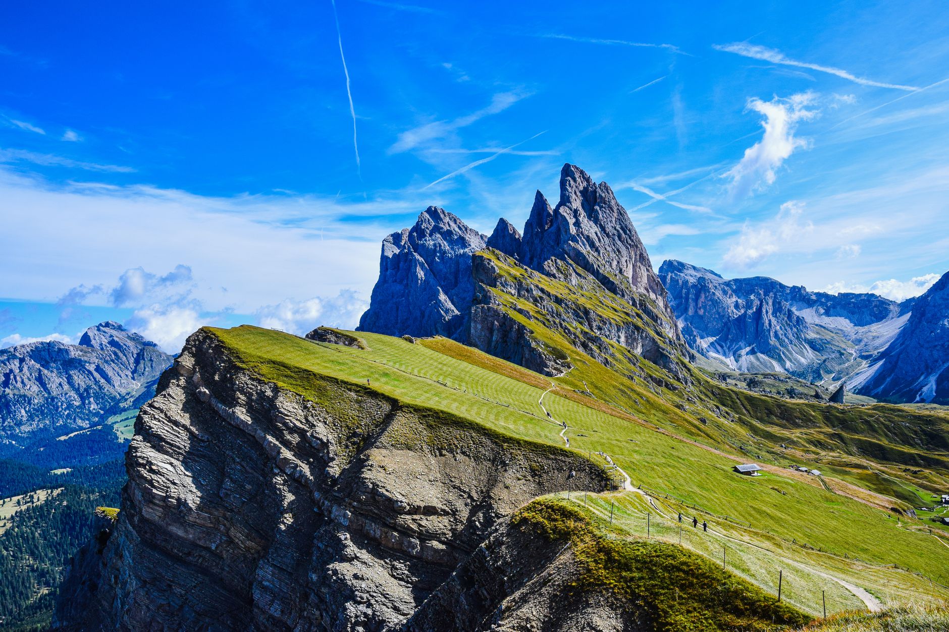 Sweeping panoramic view of the Dolomites in summer, showing jagged peaks, green alpine meadows, and a clear blue sky.