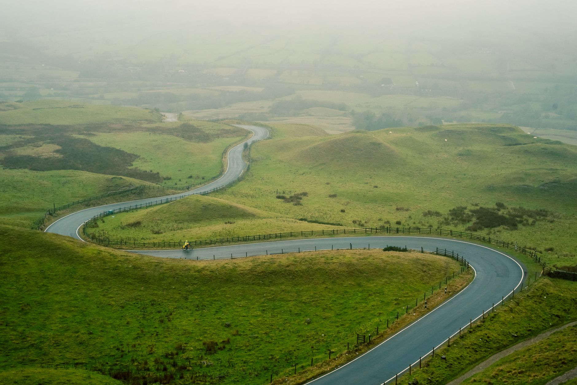 Sweeping view of a misty, ancient British countryside with rolling green hills and a hint of an old stone structure in the distance.