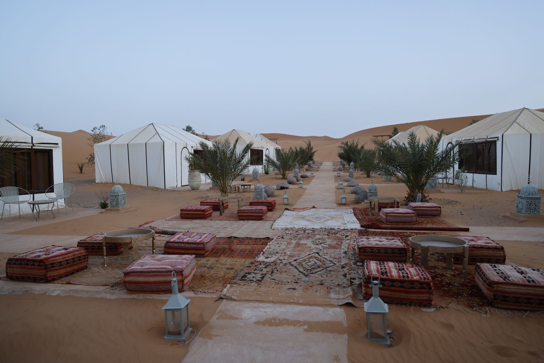 A sweeping panoramic view of a vast, golden desert landscape under a clear sky, with a traditional Bedouin tent camp nestled among the dunes and a faint caravan trail in the distance.