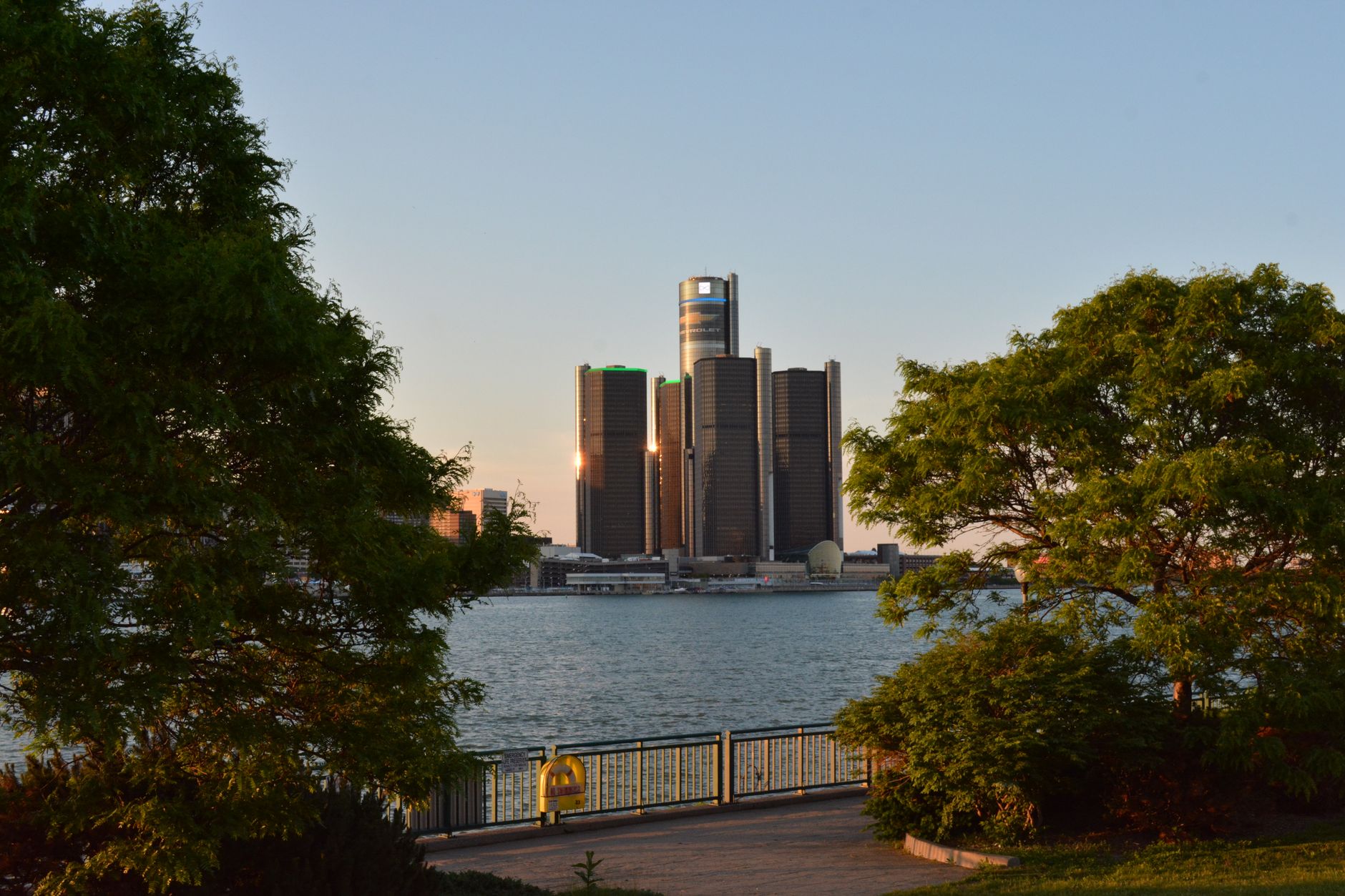 A wide, sun-drenched view of the Detroit skyline from the Detroit River, with green parks and modern architecture visible under a clear blue sky.