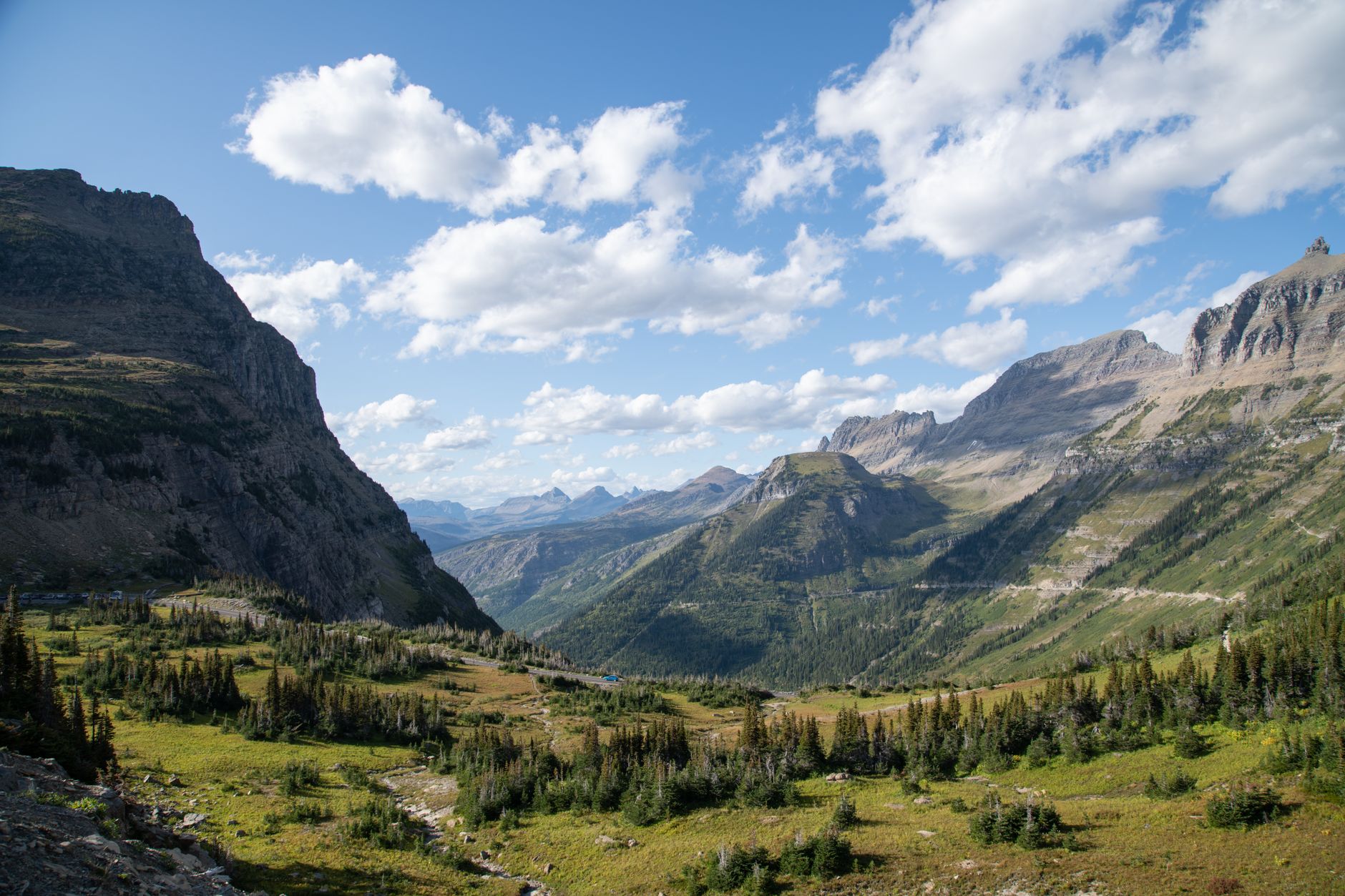 A vast, sweeping panoramic view of Glacier National Park, showing jagged, snow-capped mountains, a pristine turquoise lake, and dense pine forests under a clear blue sky.