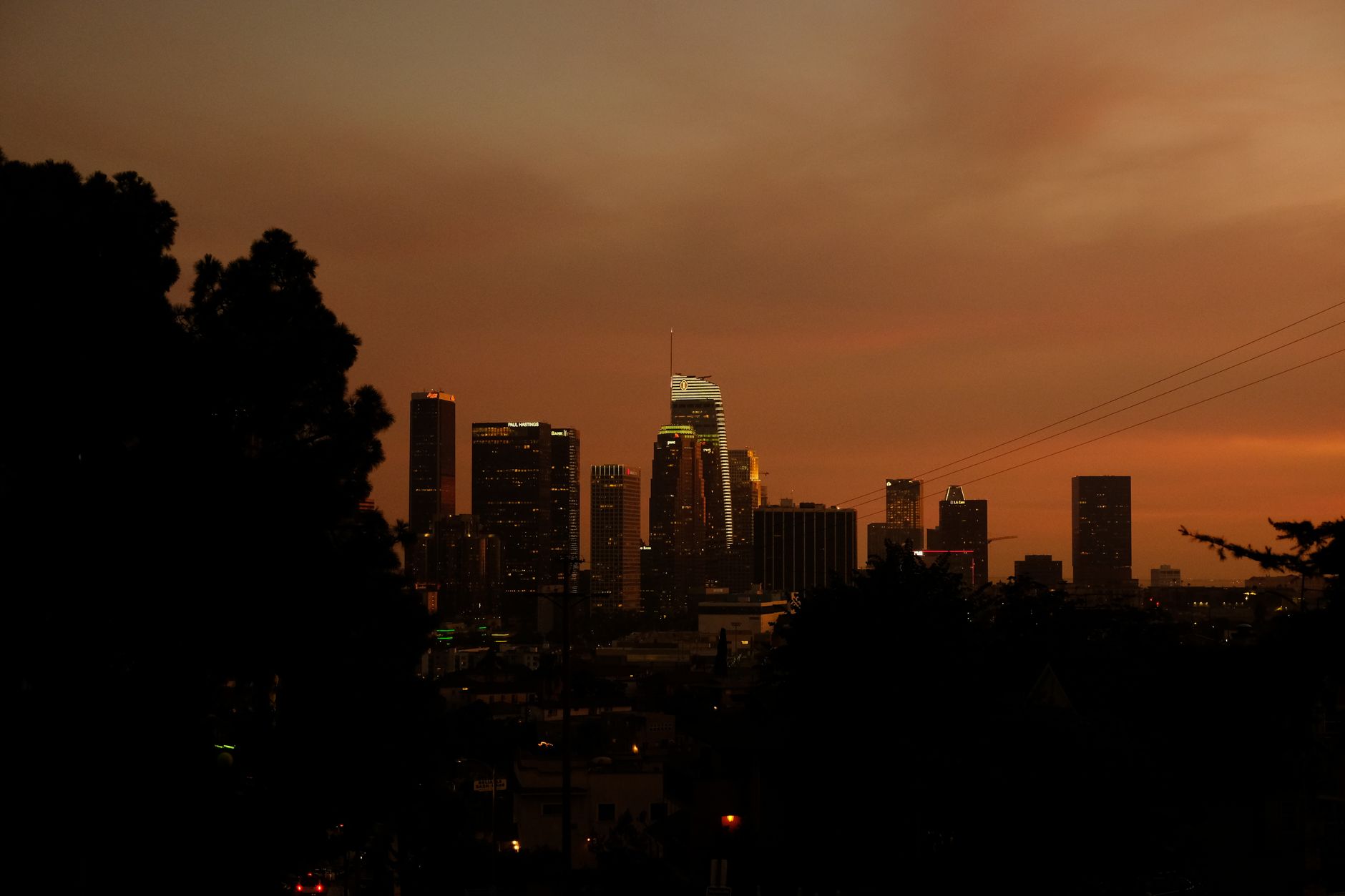 A sweeping view of the Los Angeles skyline at dusk, with pockets of urban light contrasting with dark, distant hills and an atmospheric, slightly ominous sky.