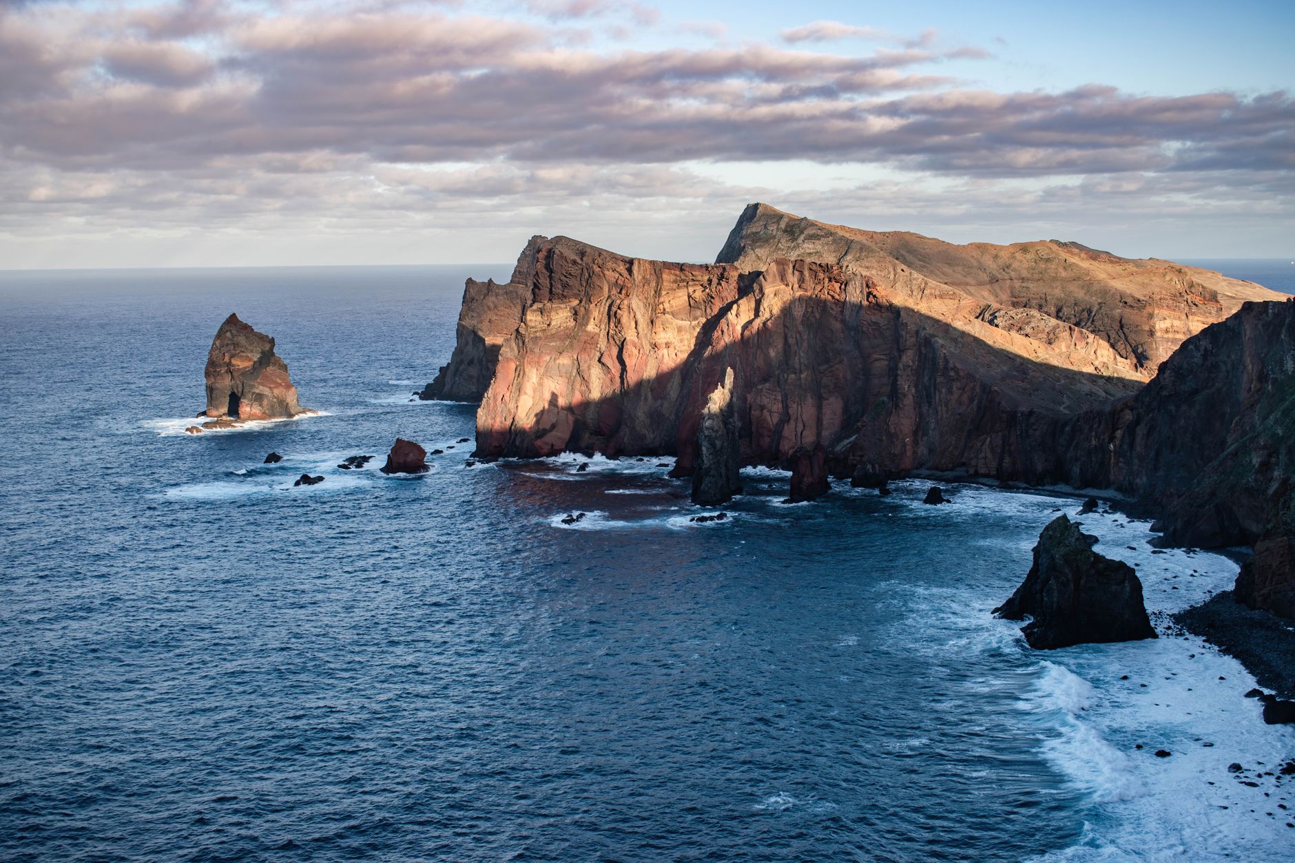 Aerial view of a rugged, wild Portuguese coastline with dramatic cliffs, golden sands, and crashing Atlantic waves under a clear sky.