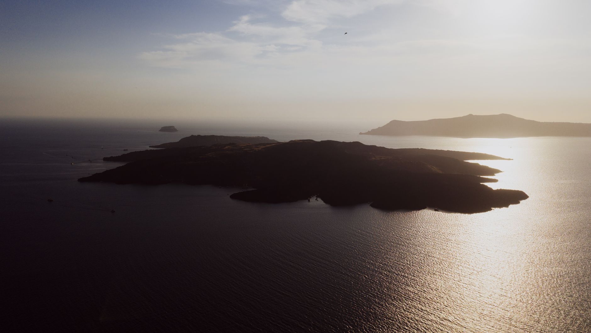 An aerial view of Santorini's unique volcanic coastline, featuring dark sand beaches contrasting with the bright blue Aegean Sea under a clear summer sky.