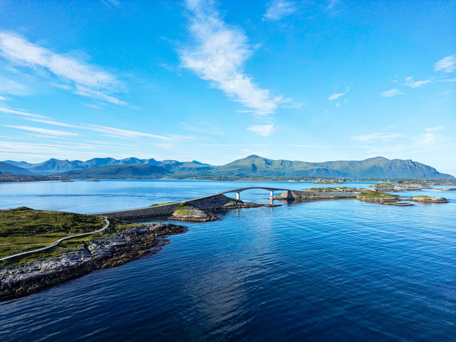 Sweeping panorama of a Norwegian fjord with steep green mountains, a blue sea, and a small village nestled on the shore under a bright summer sky.