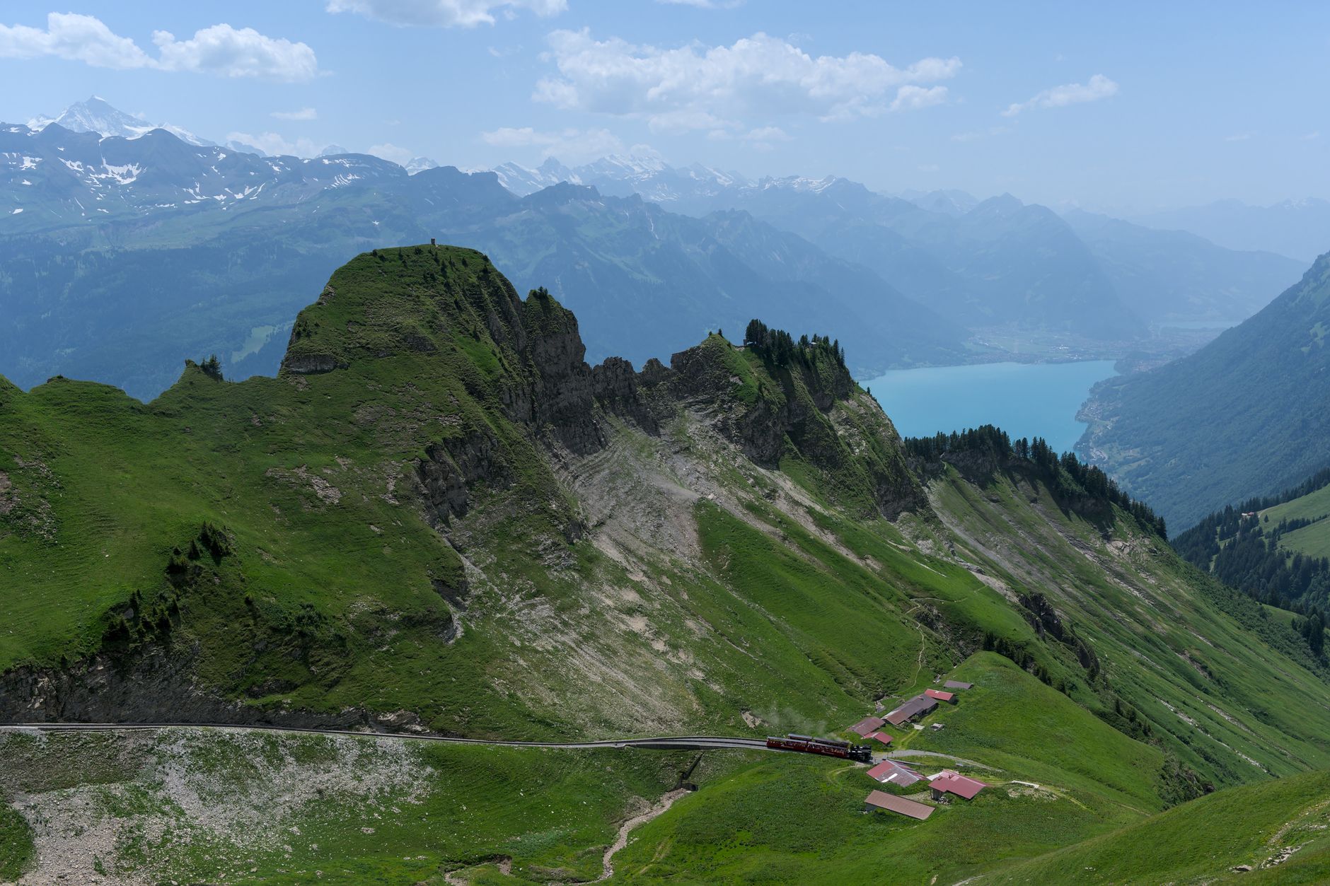 A panoramic view of a vibrant turquoise alpine lake in the Swiss Alps, surrounded by lush green mountains under a clear blue sky.