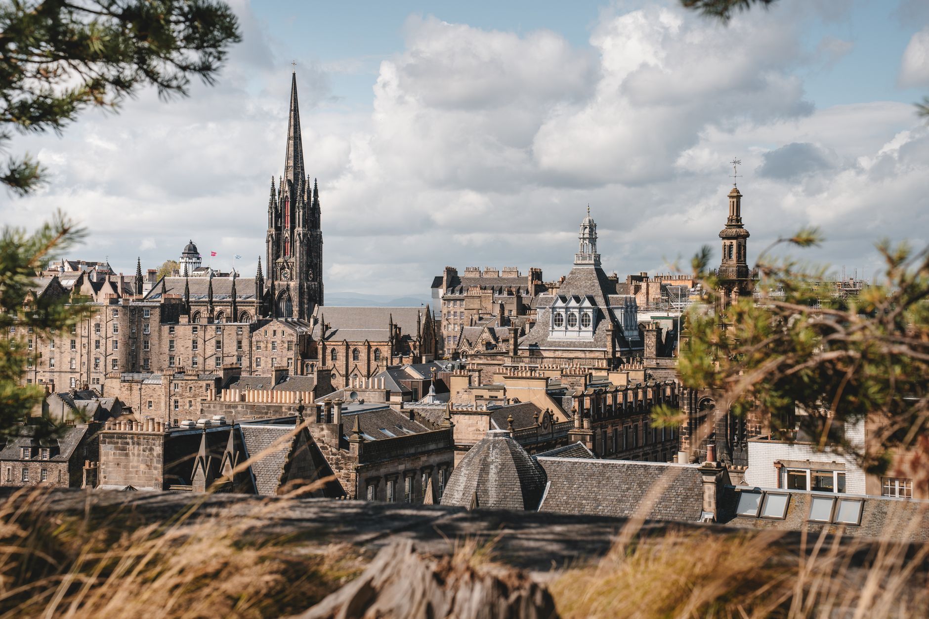 A sweeping panoramic view of Edinburgh, showcasing its historic Old Town buildings, the castle in the distance, and the green slopes of Arthur's Seat under a dramatic sky.