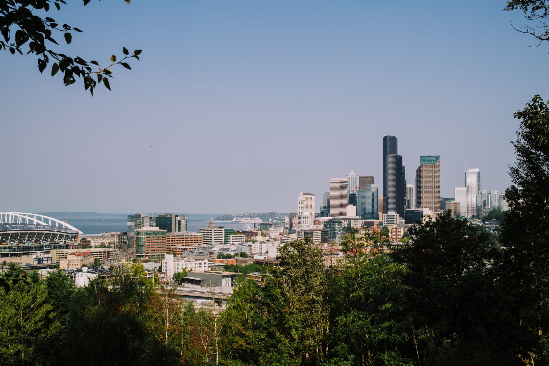 Sweeping view of Seattle's skyline at sunset from Gas Works Park, showing the city lights reflected on Lake Union with lush green hills and industrial structures in the foreground.