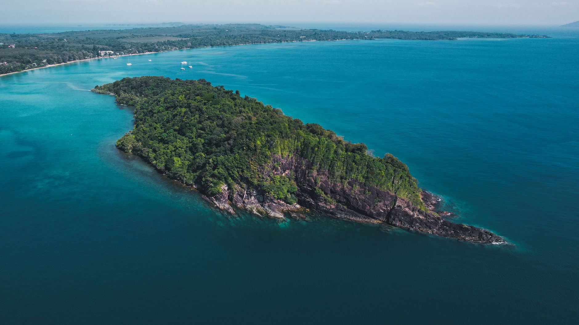 Sweeping drone view of a pristine, secluded beach in Southern Thailand, framed by lush green cliffs and turquoise water under a clear sky.