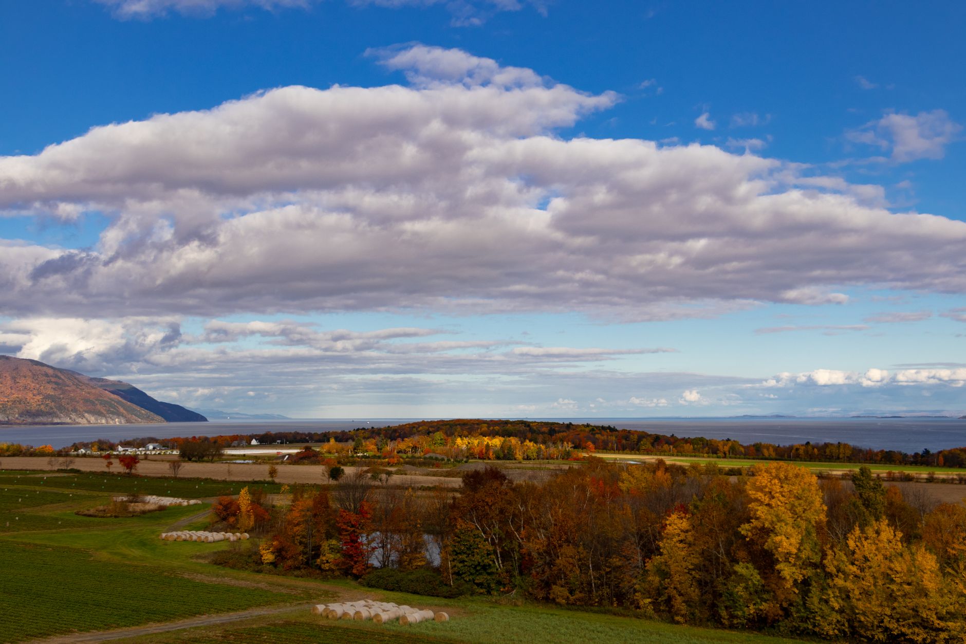 A sweeping aerial view of the St. Lawrence River winding through Quebec's vast autumnal landscape, with hints of historic architecture or a small settlement along the shore, representing the enduring French presence in Canada.