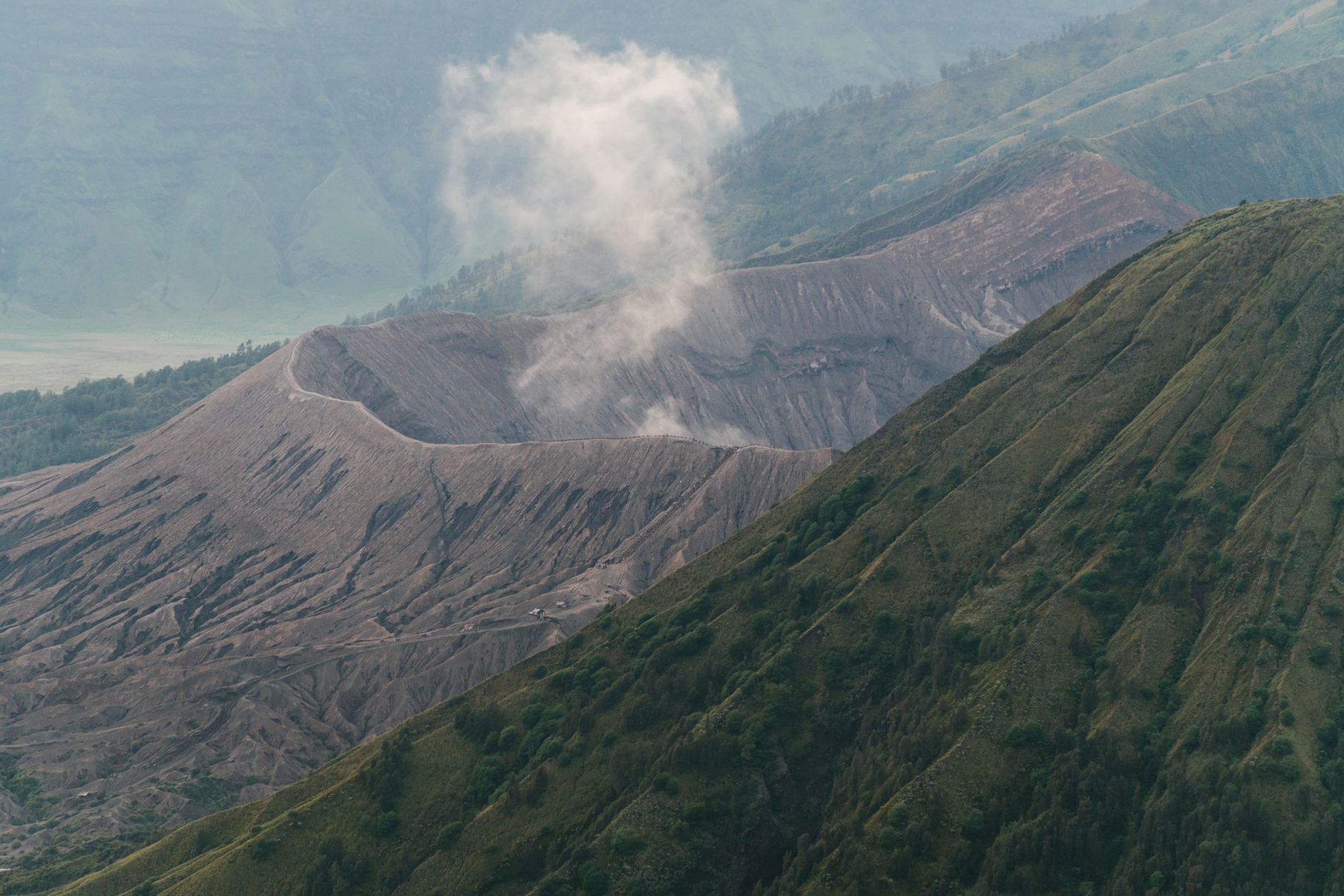 Sweeping aerial view of Mount Pinatubo's turquoise crater lake surrounded by barren, ash-covered slopes, with distant mountains under a clear sky.