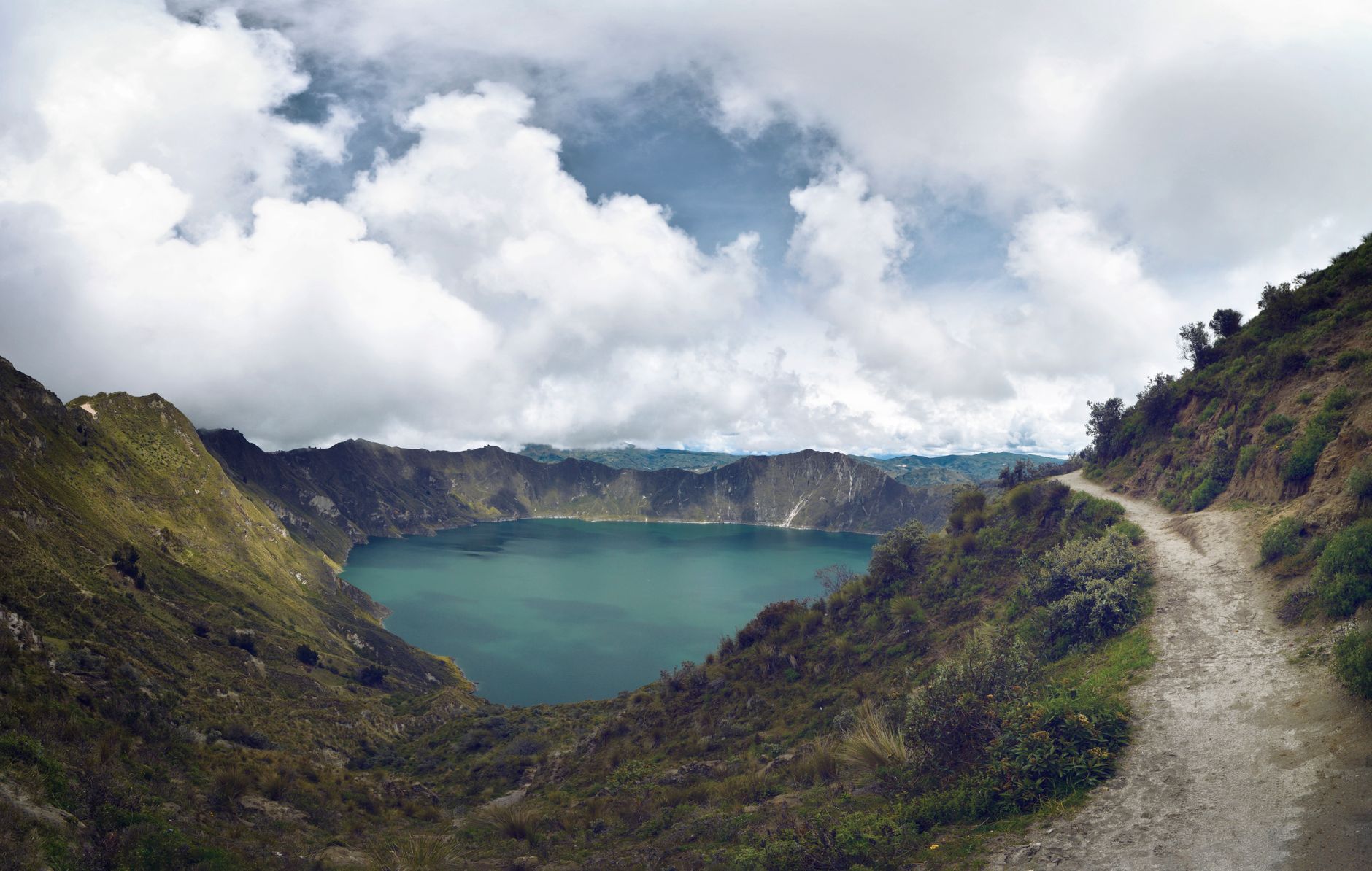 Sweeping aerial view of Mount Pinatubo's turquoise crater lake surrounded by grey volcanic ash slopes and distant green mountains under a clear sky.