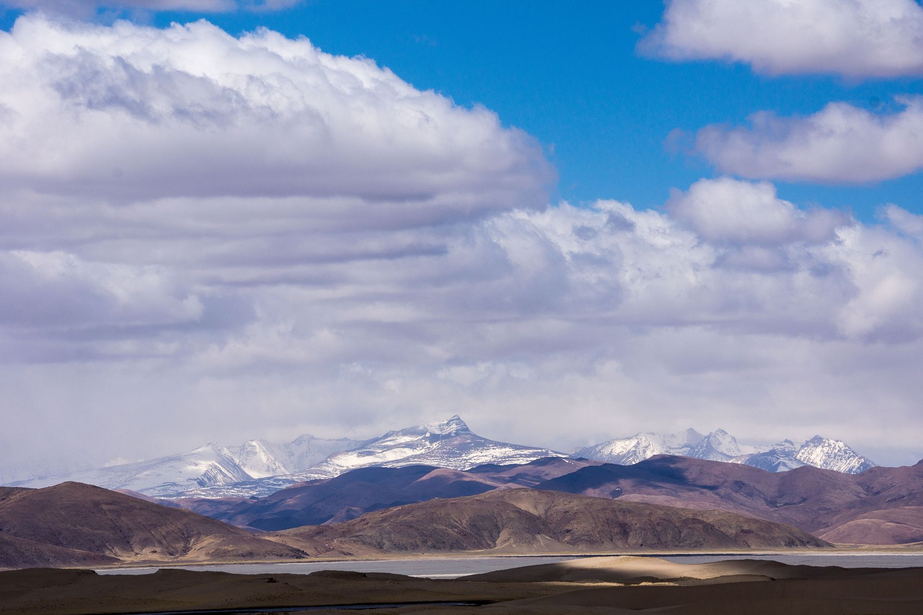 Sweeping panoramic view of a remote Tibetan village nestled in a valley, with traditional stone houses and vast mountain ranges under a clear blue sky.
