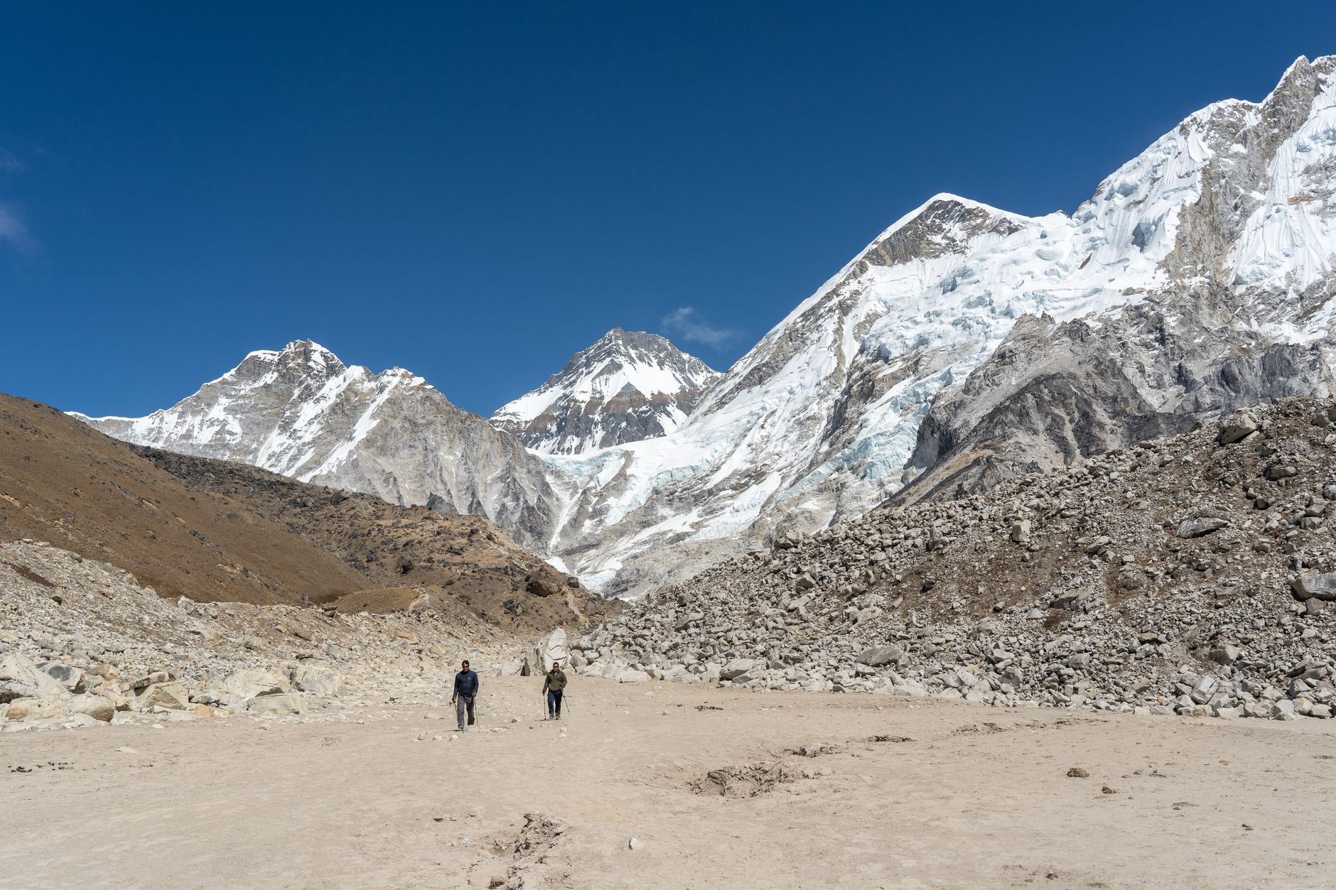 A wide aerial view of a remote Nepali village nestled in a valley, surrounded by terraced fields and towering, snow-capped Himalayan peaks under a clear blue sky.