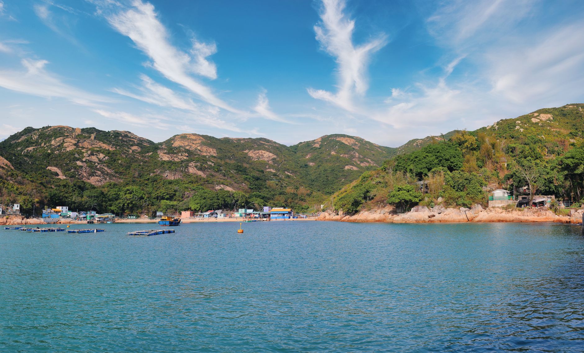 A serene view of a remote bay in Hong Kong's New Territories, with lush green hills meeting the calm sea and a small traditional village nestled on the shore.
