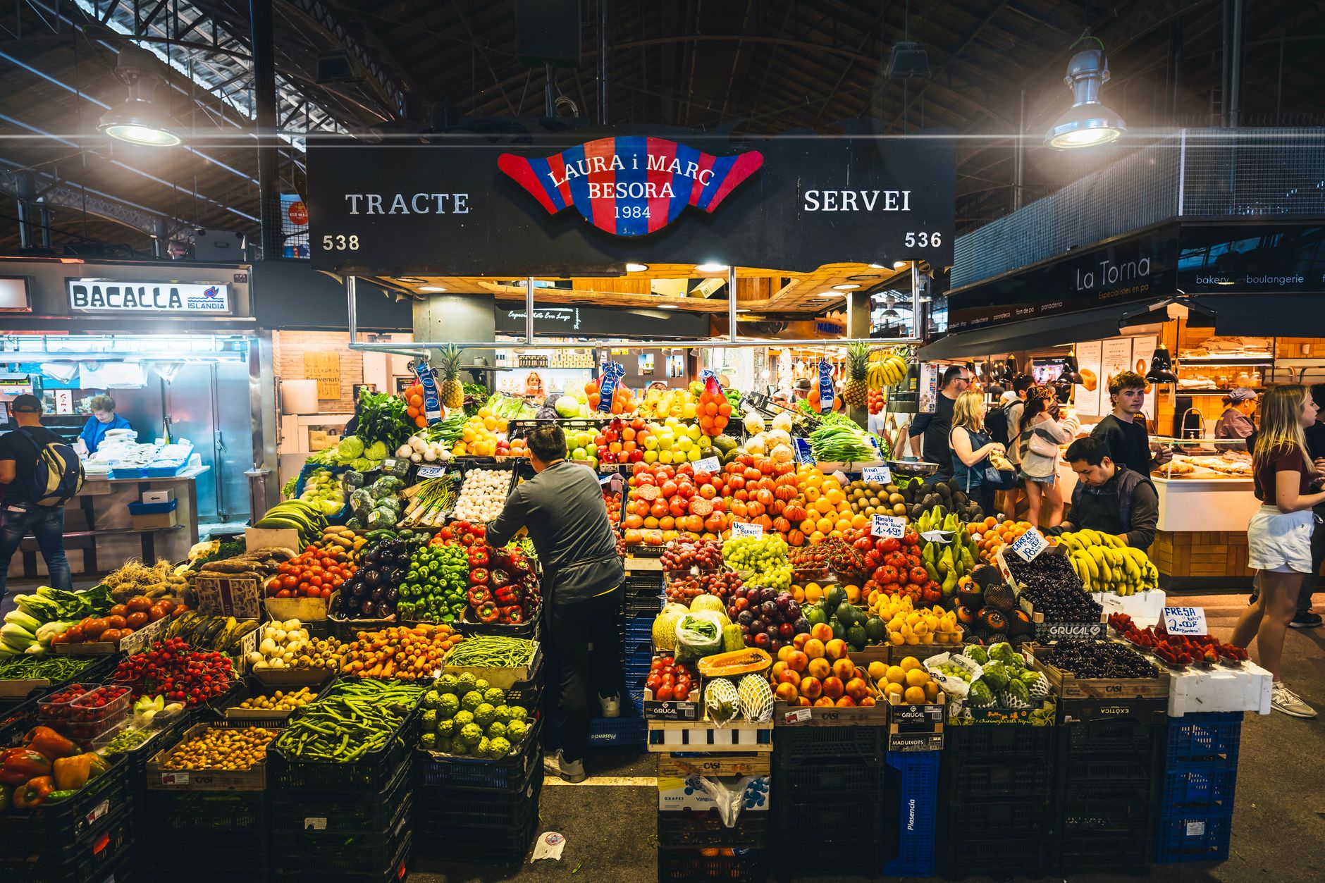 A bustling street scene in Barcelona with locals enjoying food at outdoor tables, market stalls in the background.