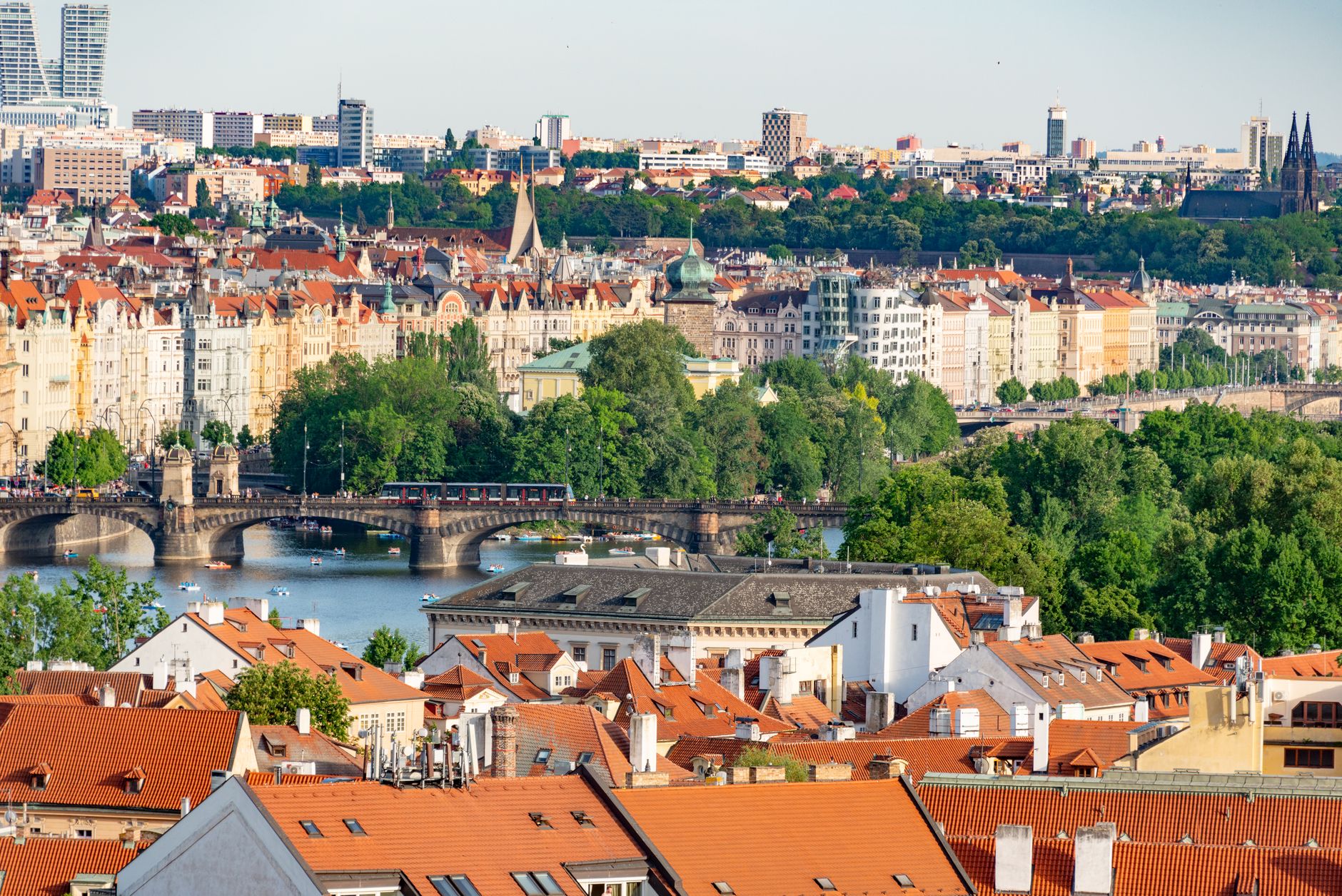 A sweeping panoramic view of Prague at sunset, showing the Vltava river winding through historic red-roofed buildings, with a vibrant market scene subtly visible in the foreground.