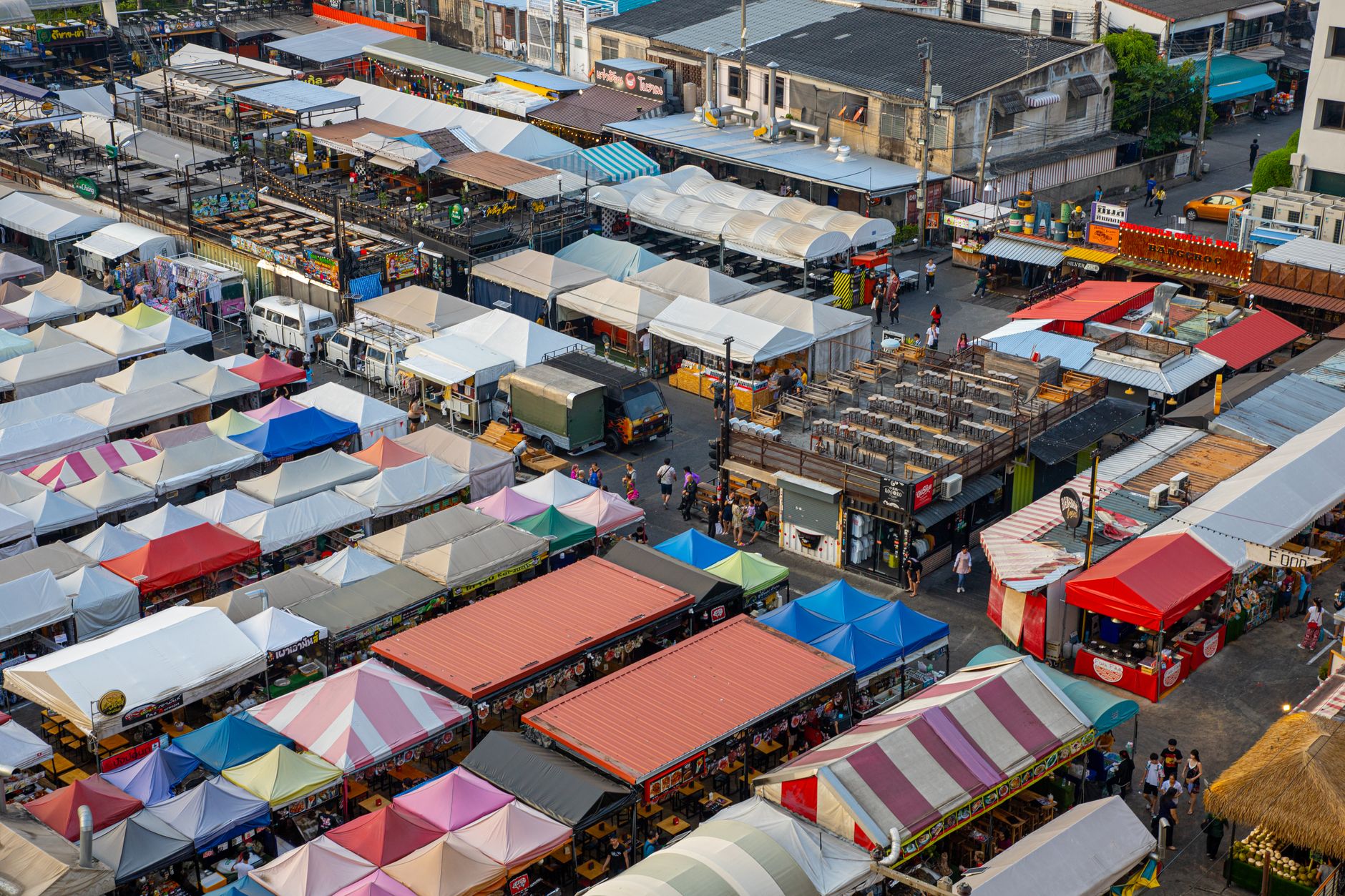 A bustling, vibrant street food market at dusk, with smoke rising from grills and colorful stalls selling various local dishes, seen from a slightly elevated perspective.