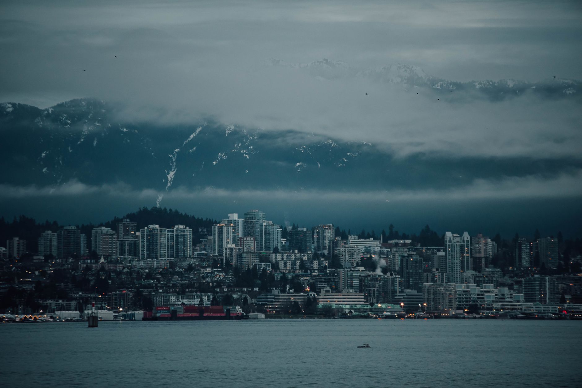 A panoramic view of Vancouver's downtown skyline nestled against the backdrop of misty coastal mountains and a calm ocean inlet, with green forests in the foreground.