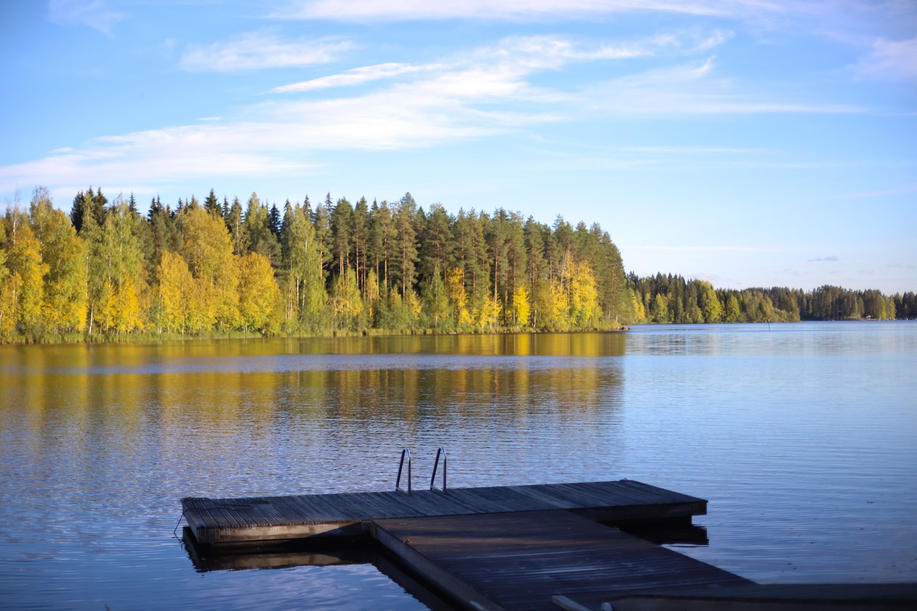 A sweeping aerial view of a serene Finnish landscape, featuring a vast, calm lake dotted with small islands, surrounded by dense evergreen forests under a soft, cloudy sky.