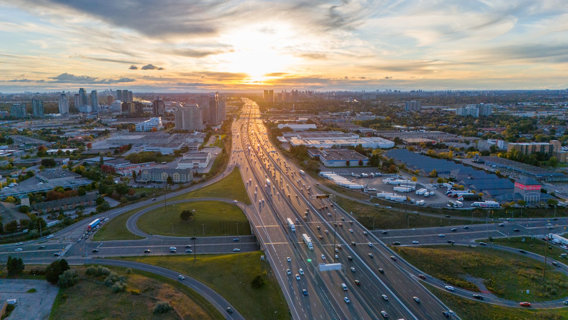An aerial view of a multi-lane highway (likely Highway 401) cutting through a green, rural landscape with scattered trees and distant water under a wide sky, illustrating the expansive, less urbanized stretch between major cities.