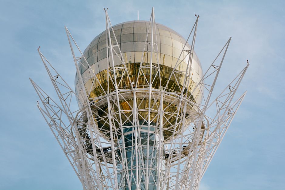 Panoramic view of the futuristic Bayterek Tower in Astana, Kazakhstan, with its golden sphere reflecting the sky, surrounded by modern architecture and a clear blue sky.