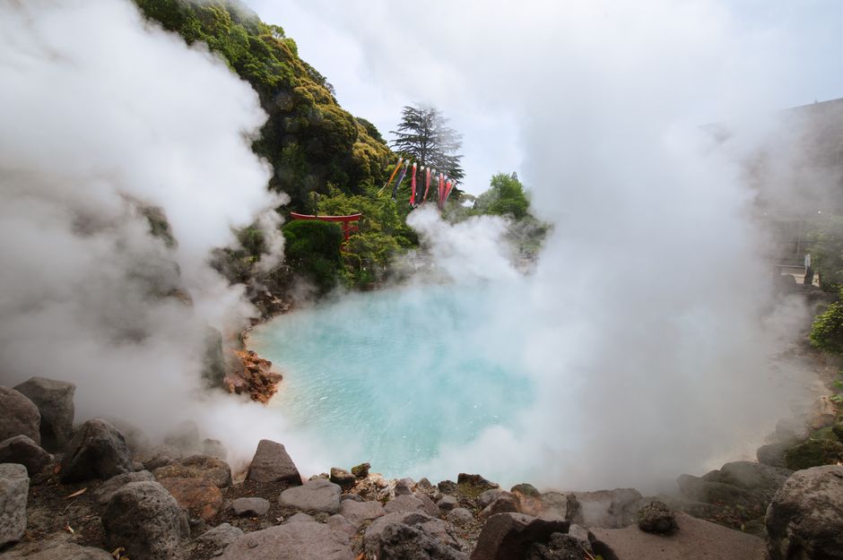 Vibrant blue Umi Jigoku hot spring in Beppu, Japan, with steam rising and lush green foliage surrounding it.
