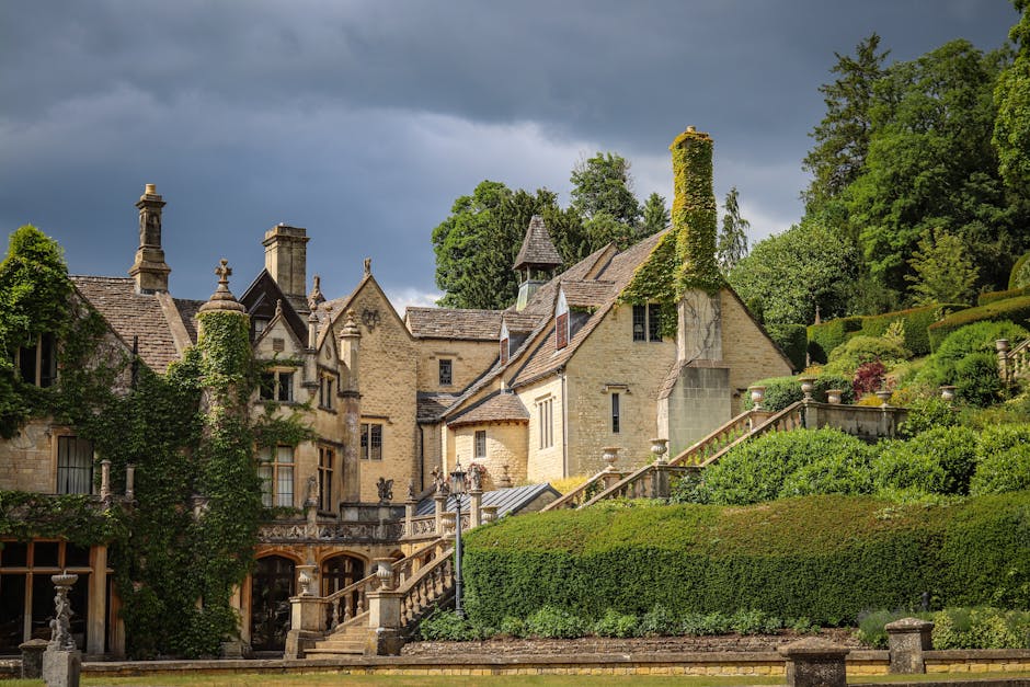 Panoramic view of Castle Combe village, England, showing historic honey-coloured Cotswold stone cottages, the Bybrook River, and St. Andrew's Church spire under a clear sky.