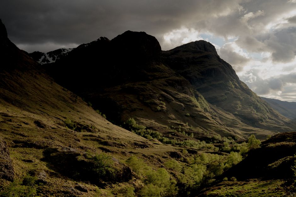 A panoramic view of Glencoe Valley in the Scottish Highlands, featuring majestic, mist-shrouded mountains, a winding river, and a narrow road beneath a dramatic sky.