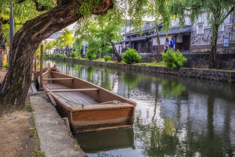 Traditional white-walled buildings line the willow-shaded Kurashiki Canal, with a flat-bottomed boat gliding on the water under a clear sky.