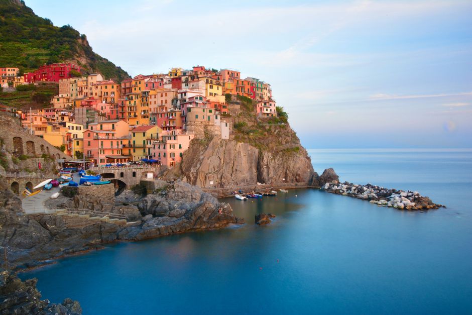 Panoramic view of Manarola, Cinque Terre, Italy, showing colorful houses cascading down a rugged cliffside towards the turquoise Ligurian Sea at sunset.