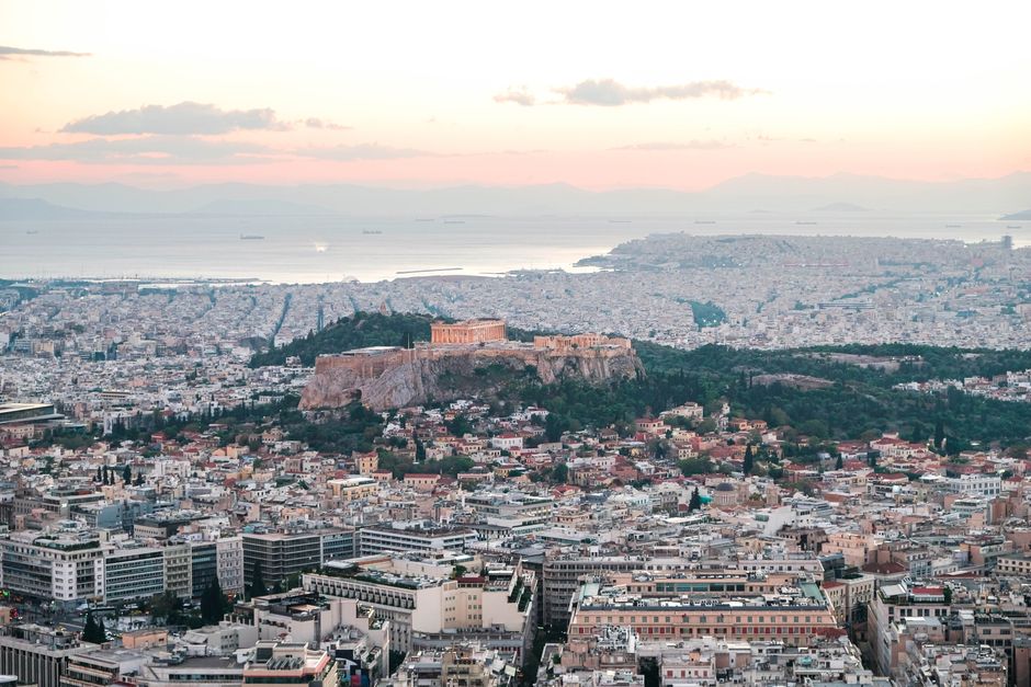 Panoramic view of Athens from Mount Lycabettus at sunset, with the whitewashed Chapel of St. George visible on the summit and the city lights beginning to glow below.