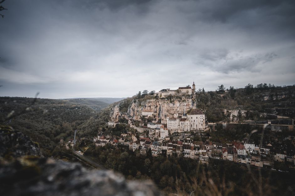 A panoramic view of Rocamadour, a medieval village built directly into a towering limestone cliff face in France, featuring stacked buildings, chapels, and a castle at its summit.